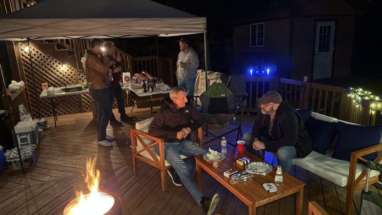 Gathering of friends on a wooden deck party at night, with a fire pit, grilling area, and socializing, captured in a cozy outdoor backyard setting.