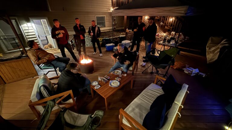 Cozy outdoor gathering around a firepit on a wooden deck with friends at night, featuring casual seating, warm lighting, and a relaxed atmosphere for socializing and celebrating.