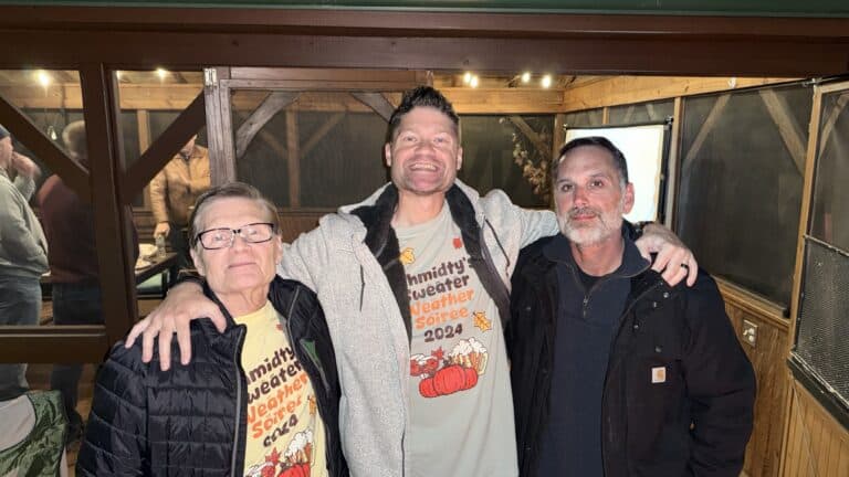 Portrait of three smiling people with arms around each other at a cozy indoor event, wearing matching themed t-shirts; celebrating autumn weather with friends and family.