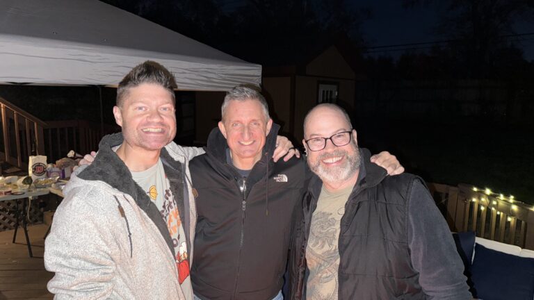 Happy men at outdoor gathering, friends enjoying evening together on wooden deck with string lights, socializing and smiling, casual night-time event, male friends friendship, outdoor party, evening socializing, revelry, fun and laughter, seasonal celebration, summer night.
