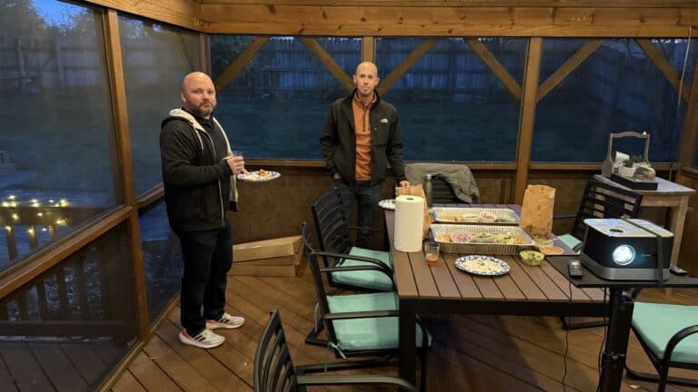 Cozy outdoor dining area with two men enjoying a casual gathering on a screened porch with food and drinks, perfect for backyard parties and outdoor entertaining, featuring wood accents and ambient lighting.
