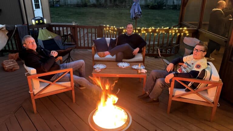 Cozy outdoor backyard gathering around a fire pit with friends during evening, featuring patio furniture, string lights, and relaxed social atmosphere.
