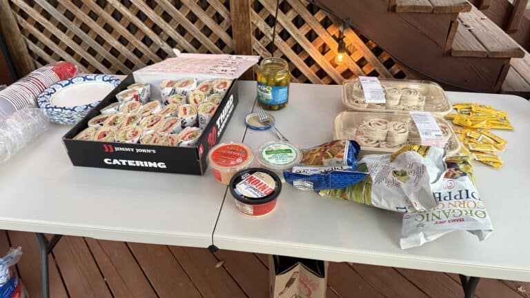 Platter with assorted pre-made sandwiches, snacks, and condiments for catering or outdoor gathering on a white folding table.